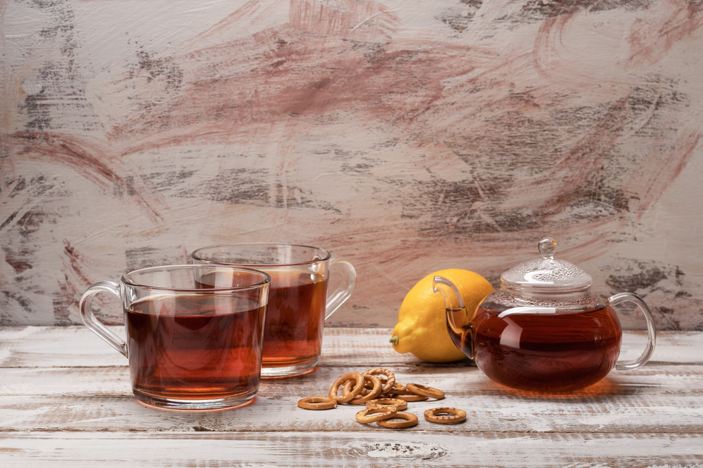 Two glass cups of reddish herbal tea beside a glass teapot, a lemon, and a rustic wooden table.