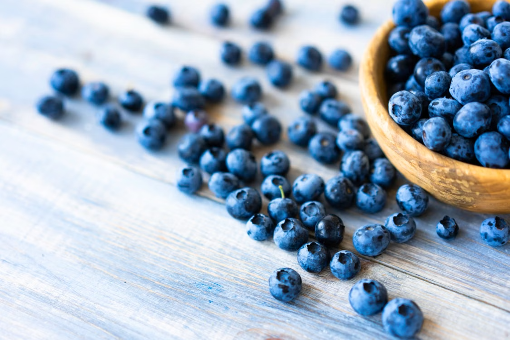 Fresh blueberries in a wooden bowl with blueberries scattered across a wooden table.